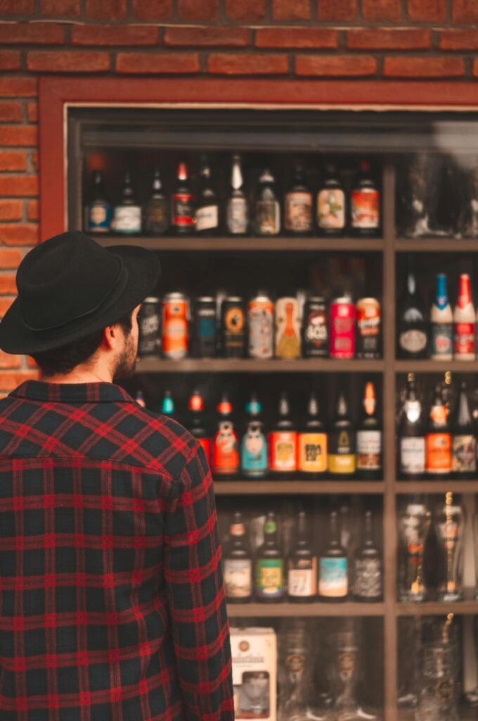 A man in a plaid shirt and hat examines a diverse beer shelf in a store.