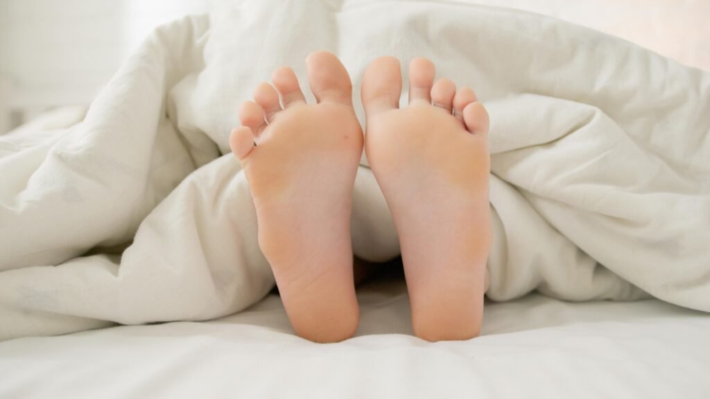 Close-up of bare feet sticking out under a soft white blanket, symbolizing comfort and relaxation.